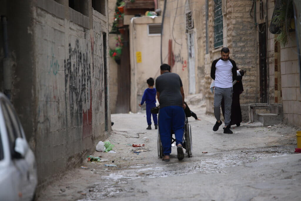 A BOY PUSHES HIS FRIEND IN A WHEELCHAIR DOWN THE STREETS OF THE CAMP. MALIK HAMAMRA/MONDOWEISS) AIDA REFUGEE CAMP, OCCUPIED WEST BANK, MAY 2023.